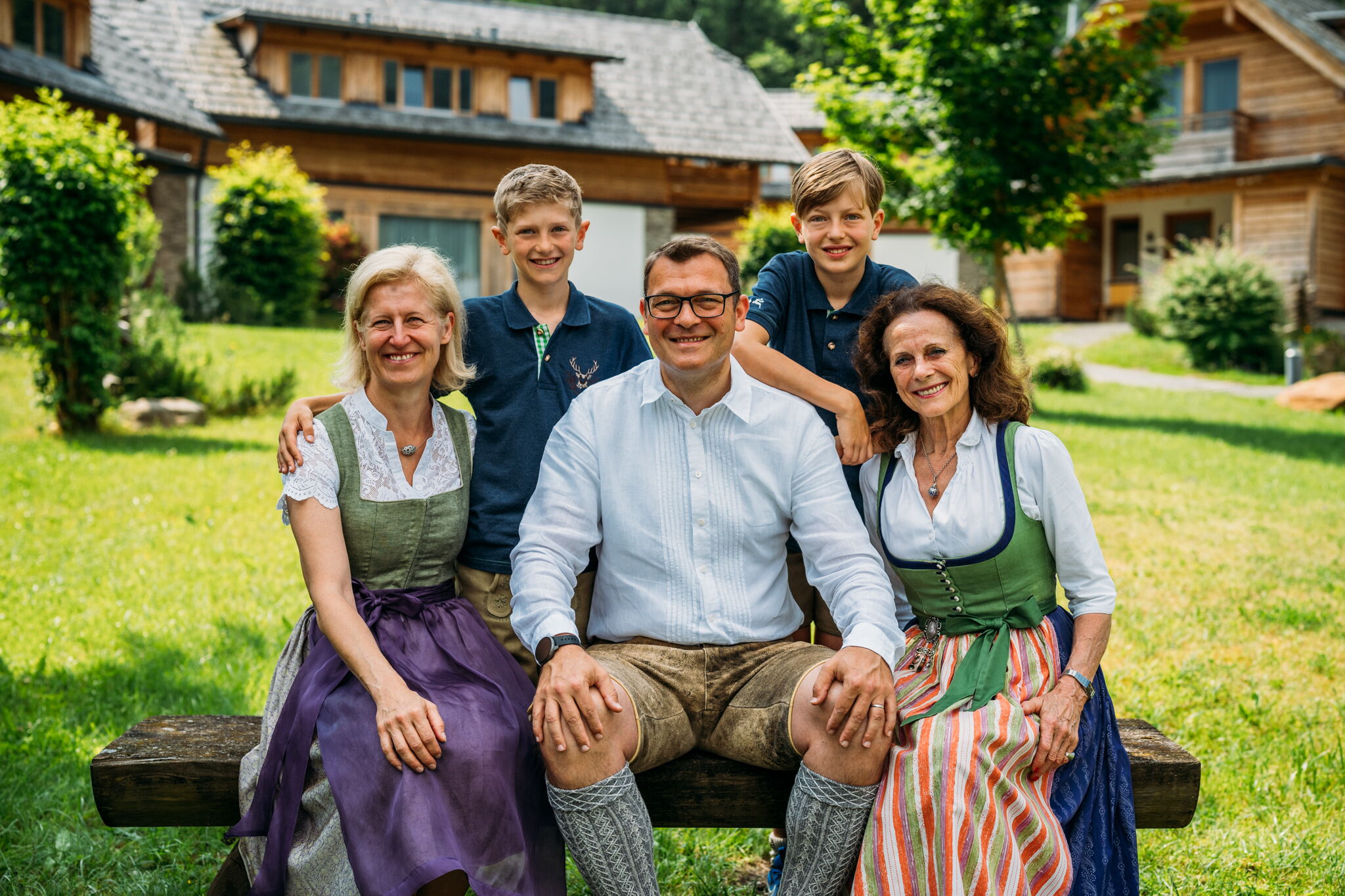 Vier Erwachsene und zwei Kinder in traditioneller Kleidung vor Holzhäusern im Grünen.