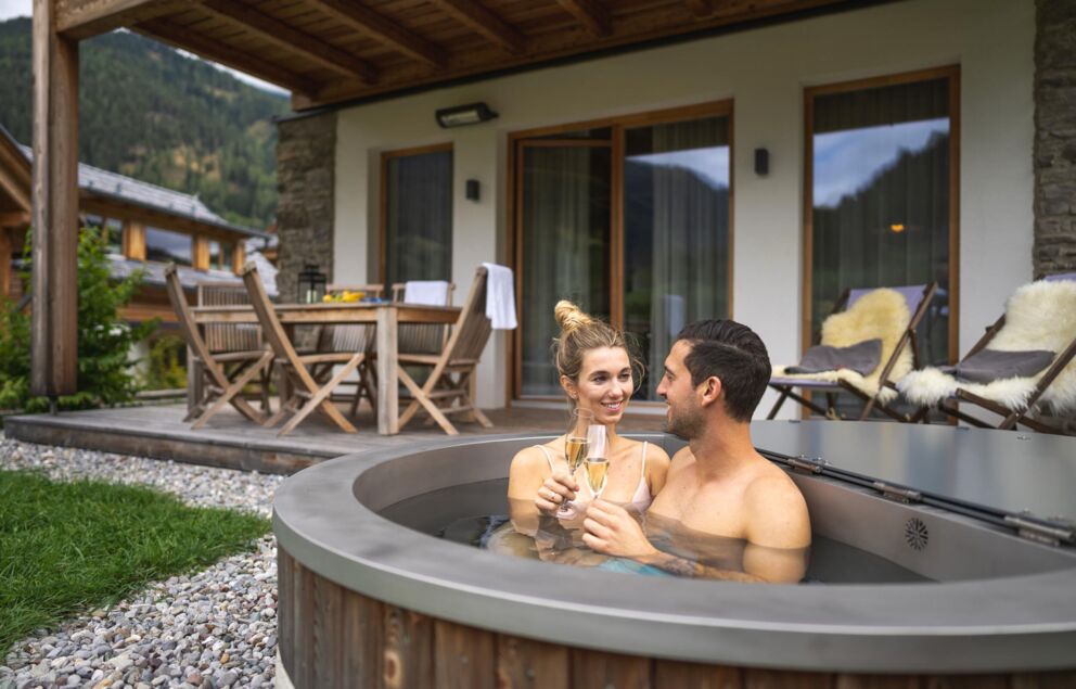 Paar im Whirlpool vor Chalet, umgeben von Natur und gemütlicher Terrasse.