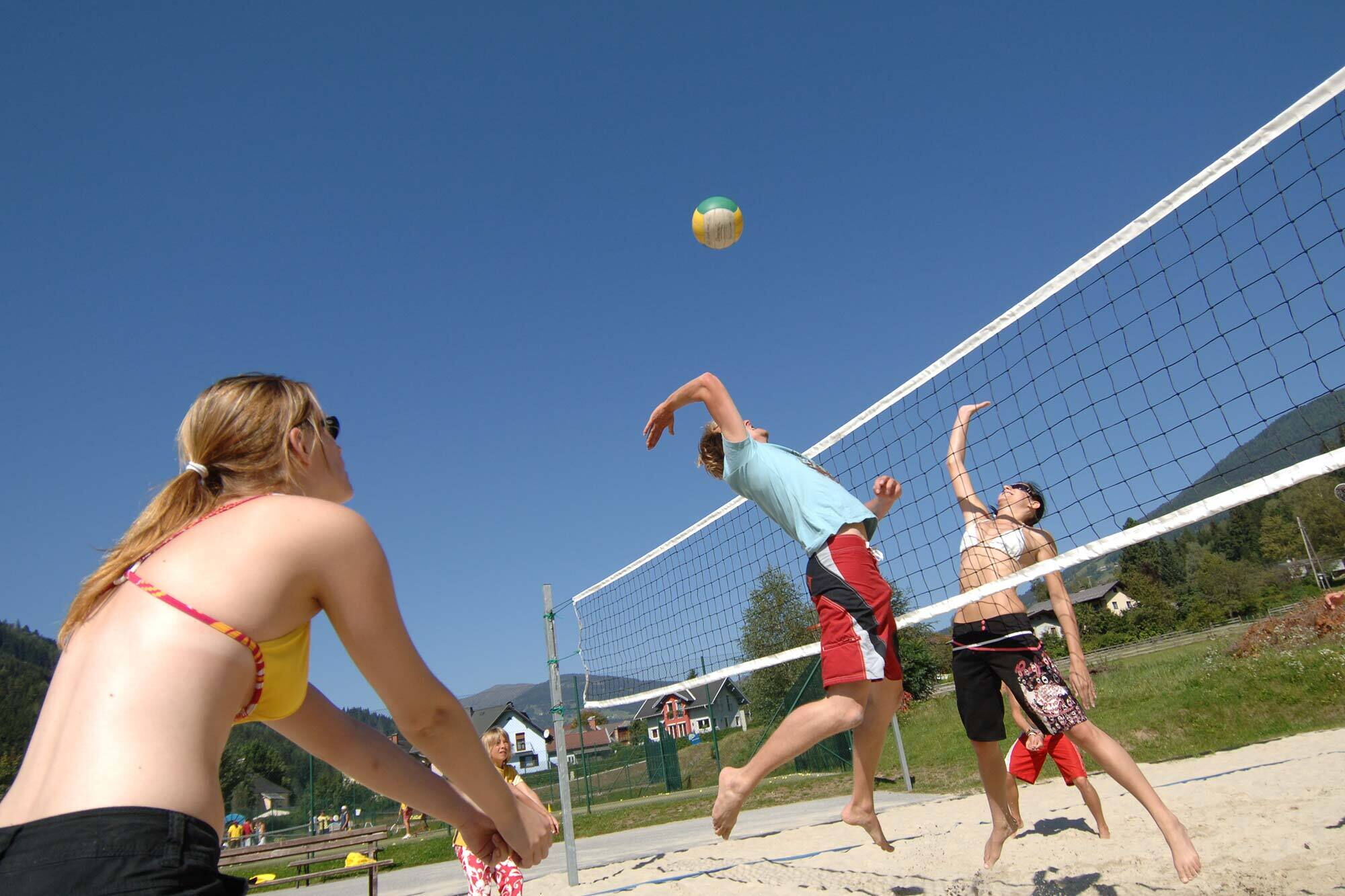 Tre persone che giocano a beach volley sotto un cielo azzurro e un sole splendente.
