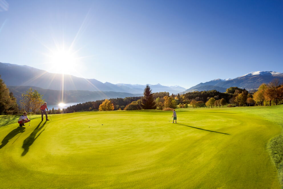 Zwei Personen spielen Golf auf einem grünen Platz mit atemberaubendem Bergpanorama.