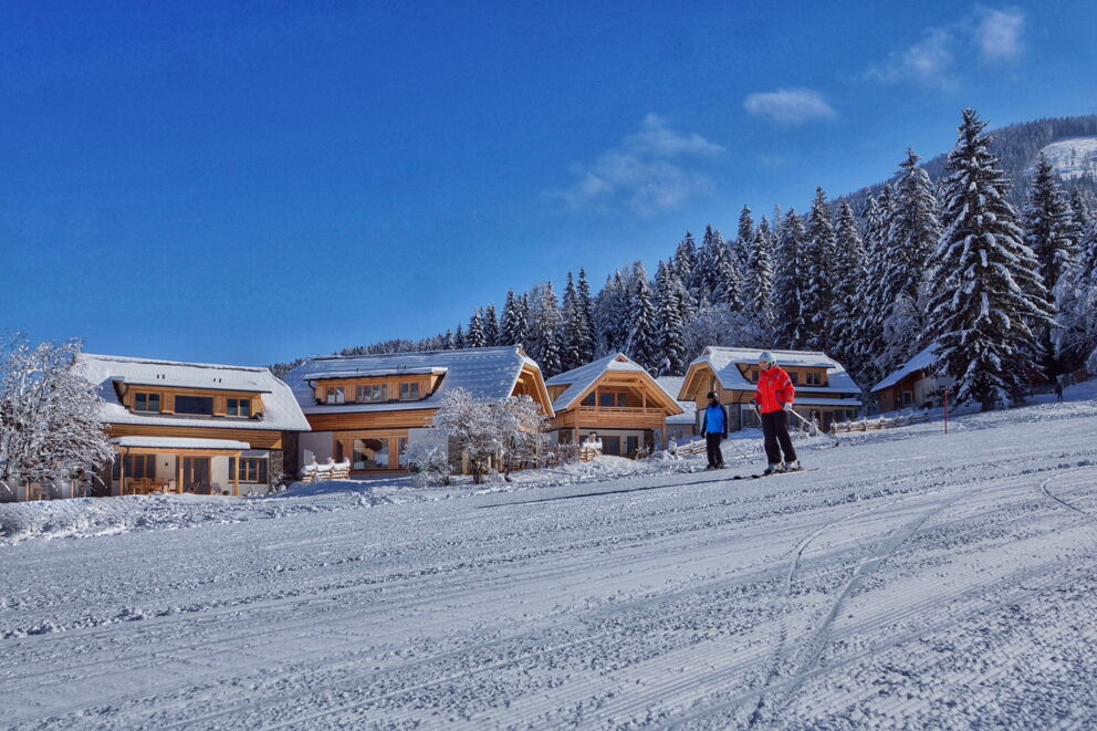 Verschneite Chalets neben Skipiste, umgeben von schneebedeckten Bäumen und strahlend blauem Himmel.