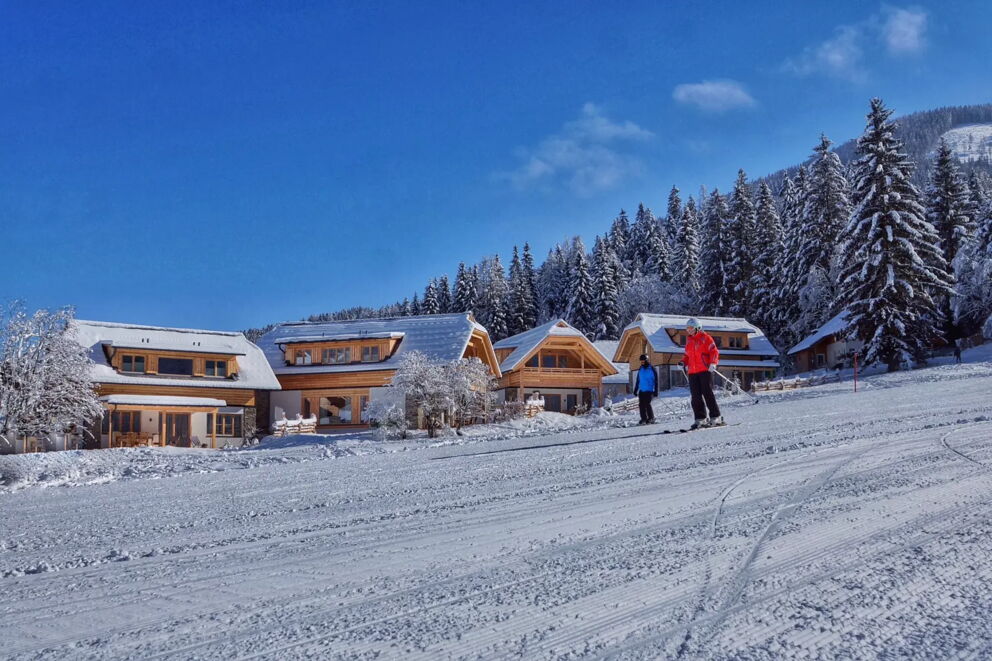 Verschneite Chalets vor majestätischem Wald und Skifahrer auf frisch präparierter Piste.