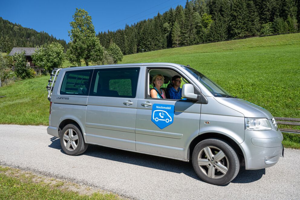 Silberner Shuttlebus auf sonniger Landstraße in grüner, bewaldeter Umgebung.