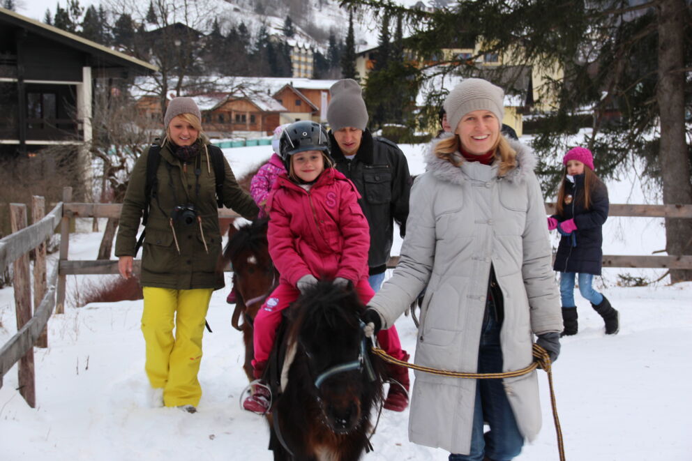 Kinder reiten Ponys im verschneiten Kärntner Winter, begleitet von Erwachsenen.