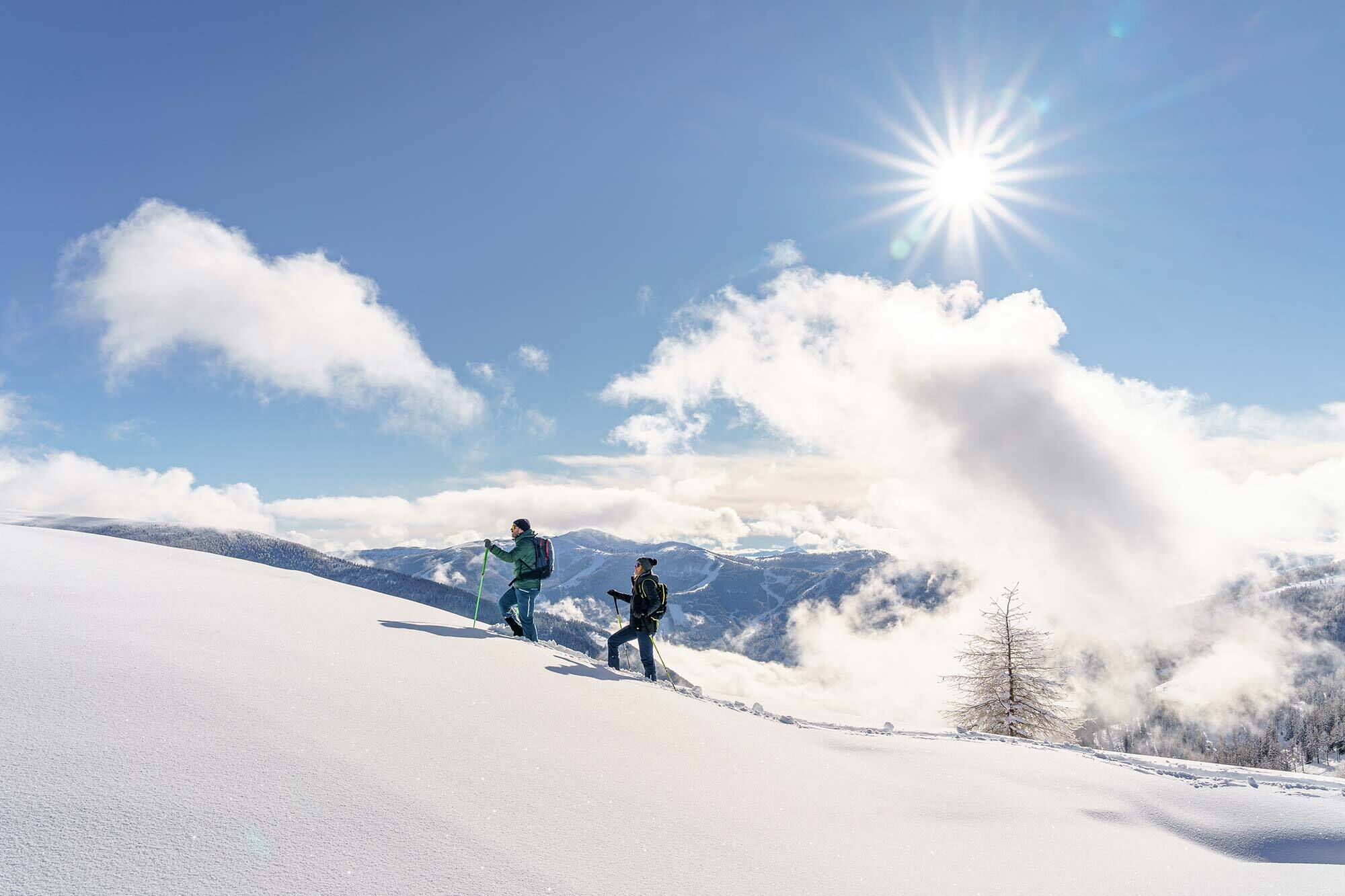 Due persone che camminano con le racchette da neve tra le montagne innevate sotto un sole splendente.