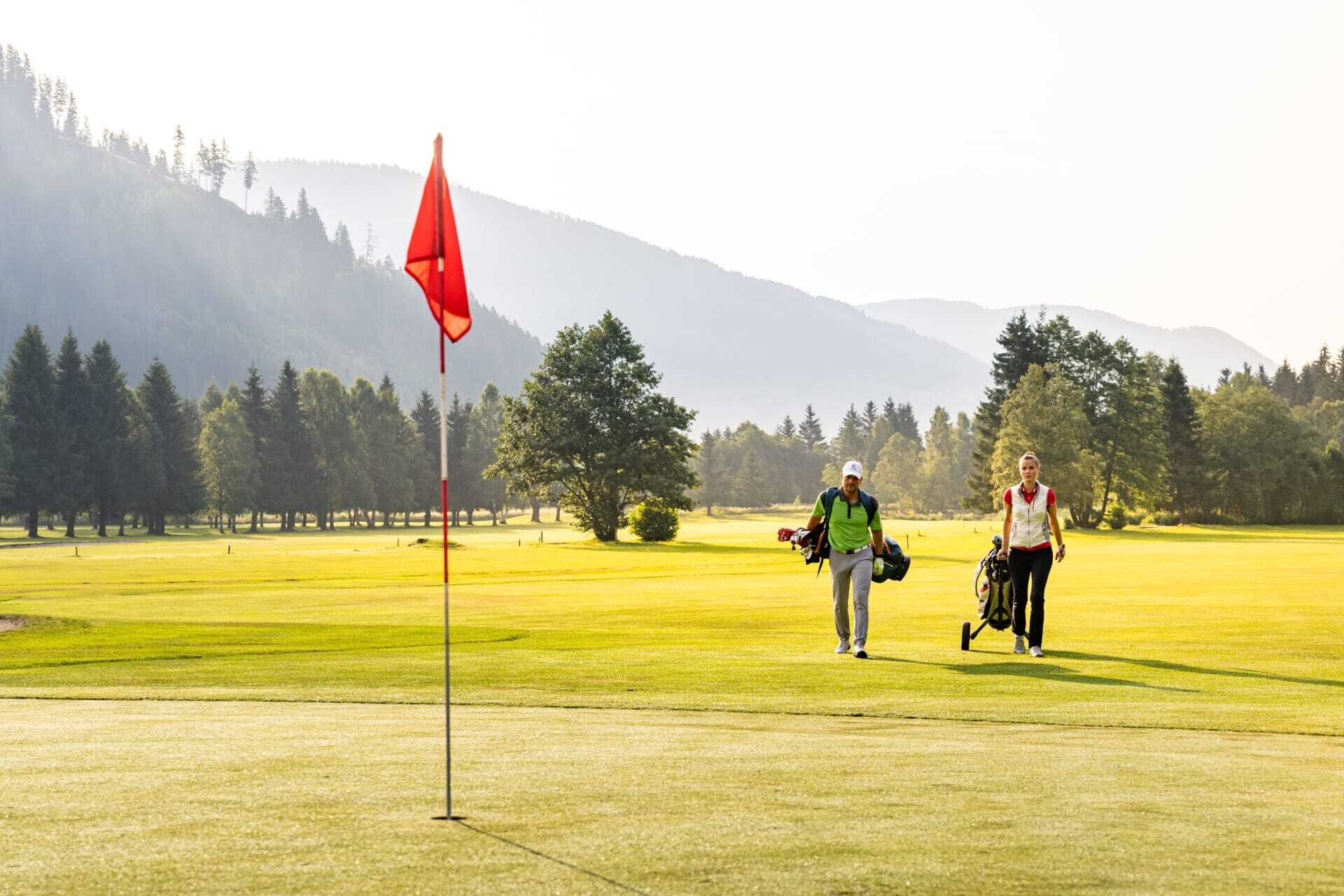 Zwei Golfspieler auf grünem Platz, umgeben von Bergen und Bäumen.