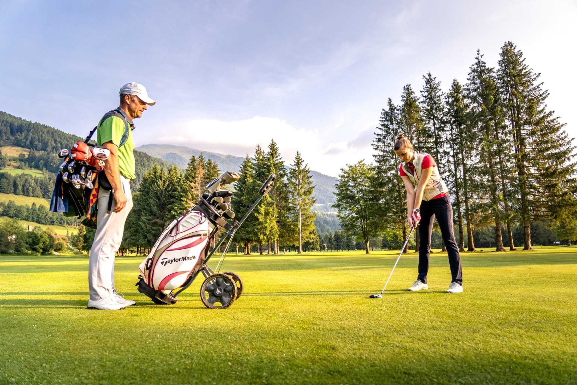 Two people play golf on a sunny course in the middle of a picturesque mountain landscape.