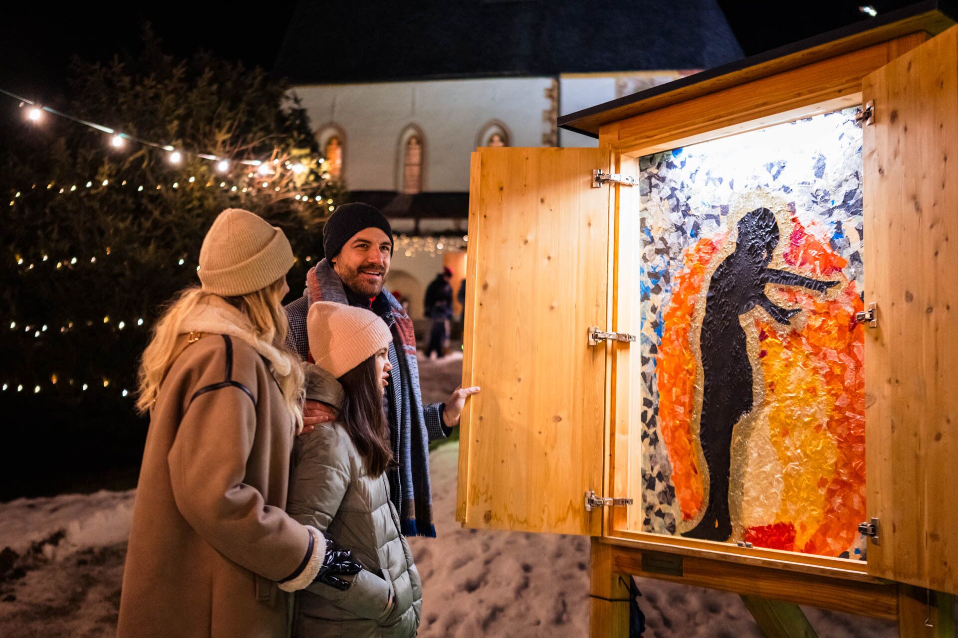 Famiglia che osserva le opere d'arte illuminate al mercato invernale, circondata da luci festose.