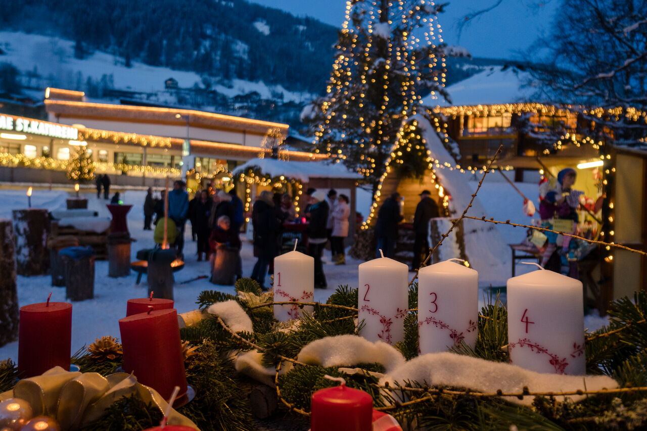 Adventskranz mit Kerzen vor festlich beleuchtetem Dorfplatz in winterlicher Atmosphäre.