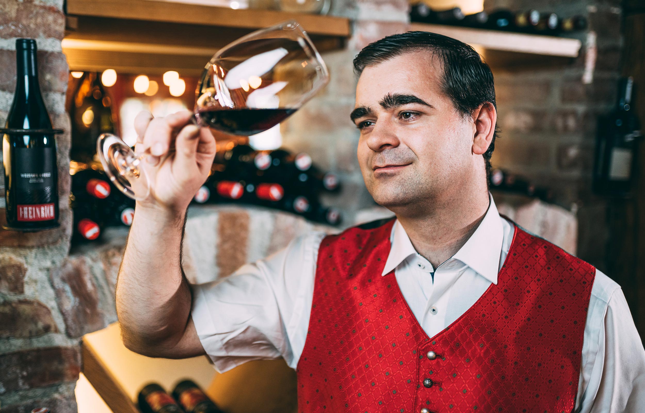 Sommelier standing in a wine cellar in front of a wine glass with red wine in it.