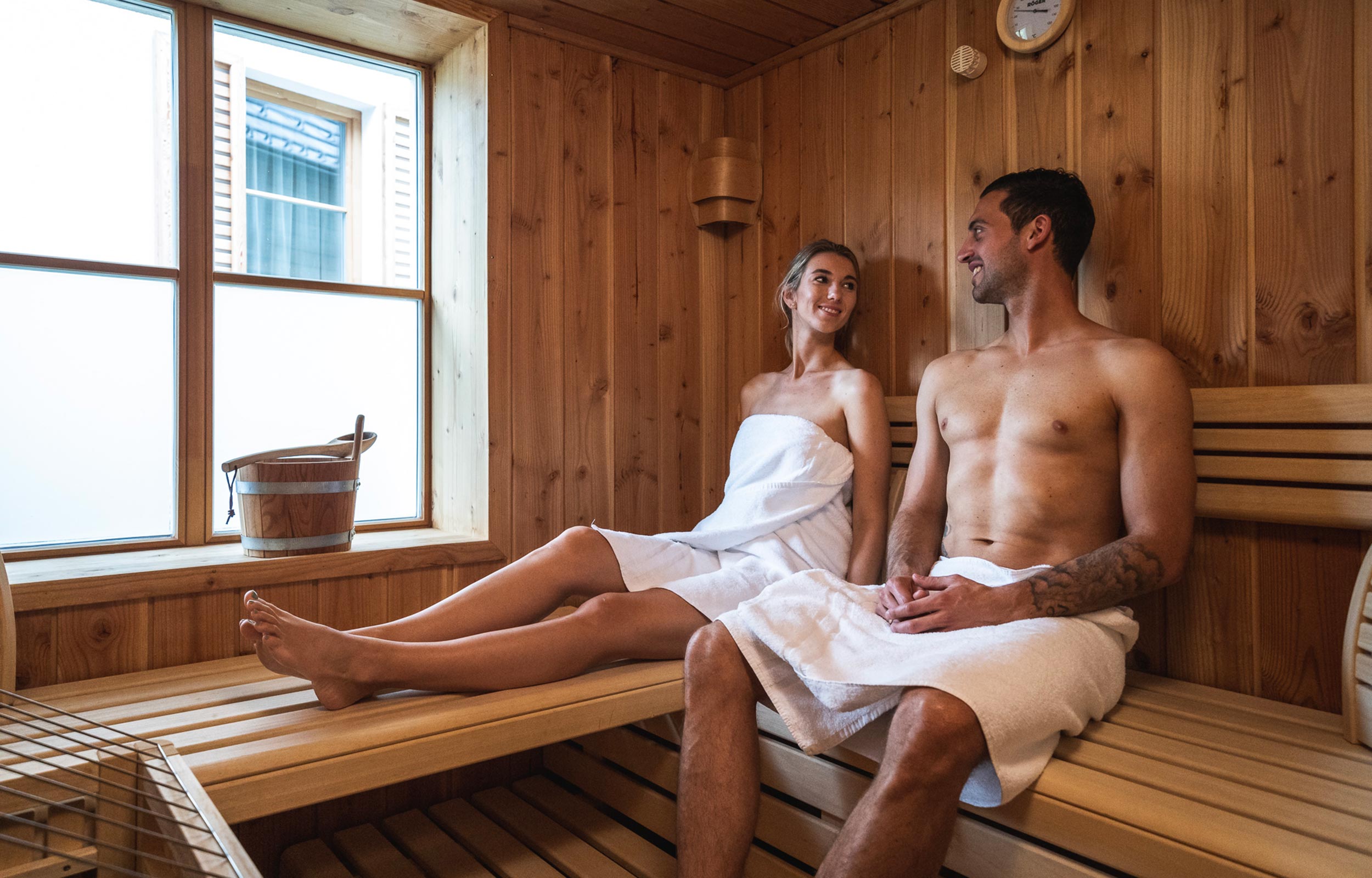 Due persone si rilassano in una sauna rivestita in legno con un'ampia finestra.