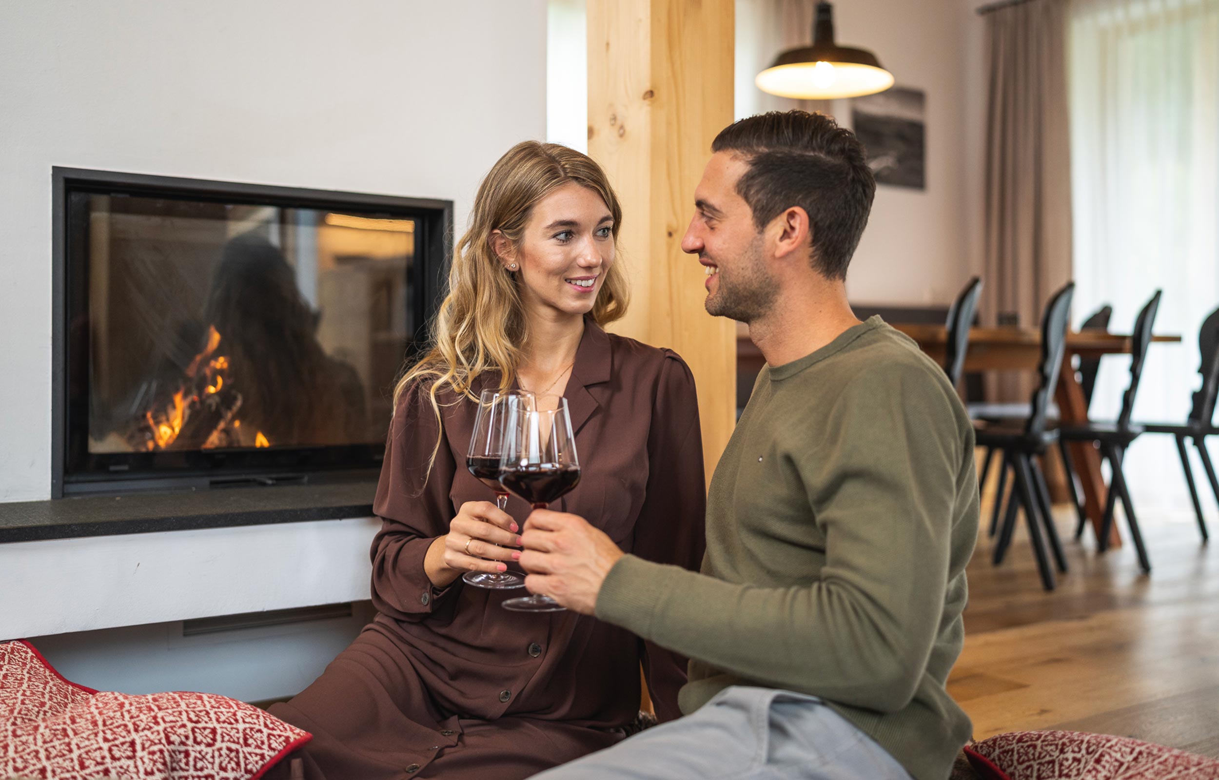 Ein Paar das romantisch vor den Kamin im Trattlers Hof-Chalets sitzt und mit Wein anstößt.