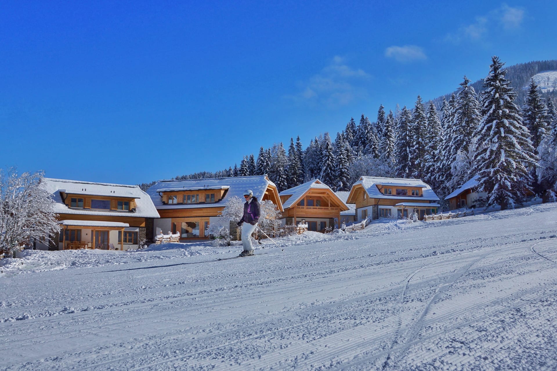Snow-covered chalets in a picturesque winter landscape with a bright blue sky and skiers.