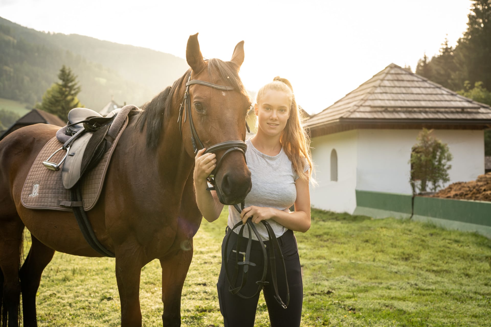 Donna con cavallo al tramonto su un prato verde davanti a uno chalet.